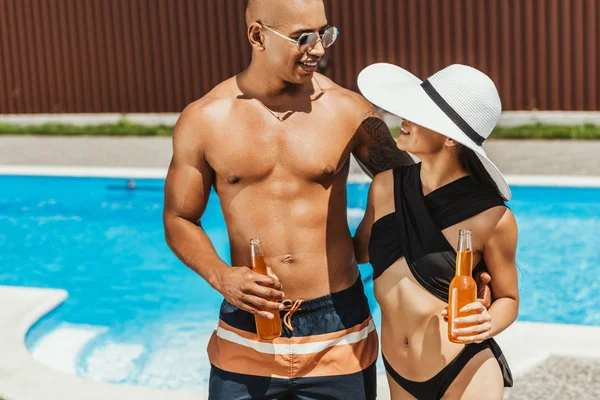 Multicultural couple holding bottles of beer and hugging at poolside — Stock Photo