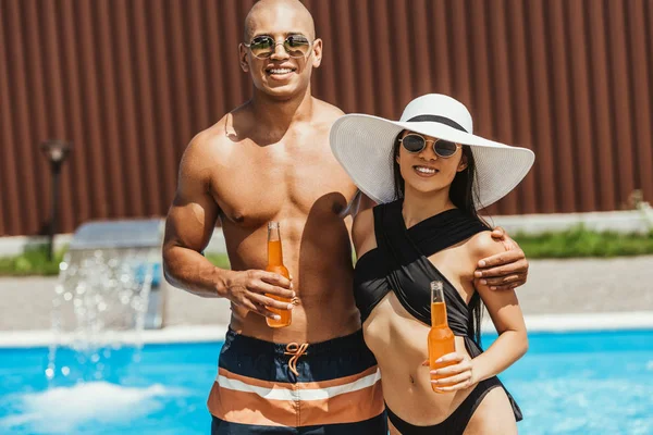 Interracial couple holding bottles of beer and embracing at poolside — Stock Photo