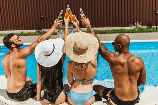Vista posteriore di amici multiculturali in costume da bagno clinking con bottiglie di birra vicino alla piscina — Foto stock