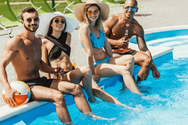 Multicultural friends with bottles of beer and beach ball relaxing at swimming pool — Stock Photo