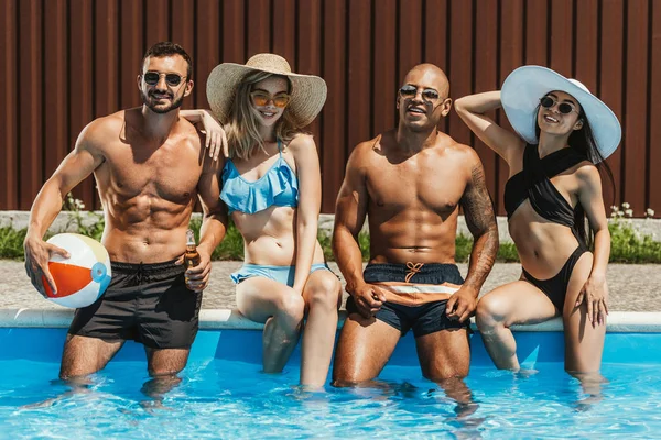 Amigos multiculturales en trajes de baño y gafas de sol sentados junto a la piscina - foto de stock