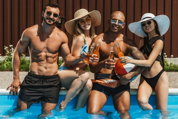 Young happy multicultural friends clinking with bottles of beer in swimming pool — Stock Photo