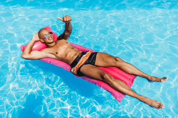 Hombre afroamericano mostrando el pulgar hacia arriba mientras toma el sol en colchón inflable en piscina - foto de stock