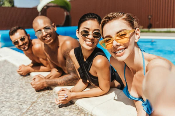Jóvenes sonrientes multiculturales en trajes de baño y gafas de sol posando en la piscina - foto de stock