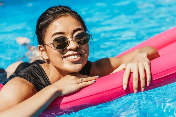 Happy beautiful asian girl in sunglasses relaxing on inflatable mattress in pool — Stock Photo