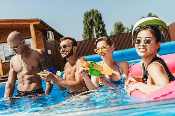 Sorrindo amigos multiculturais se divertindo com armas de água na piscina — Fotografia de Stock
