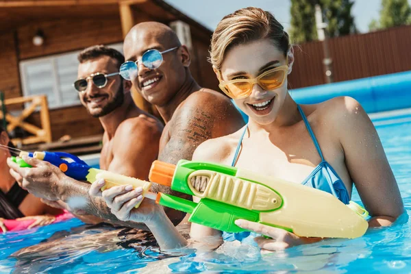 Amigos multiculturais em óculos de sol brincando com armas de água na piscina — Fotografia de Stock