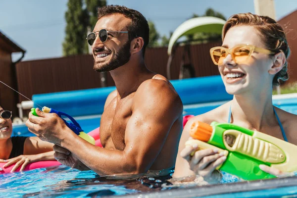 Pareja en gafas de sol jugando con pistolas de agua en la piscina - foto de stock