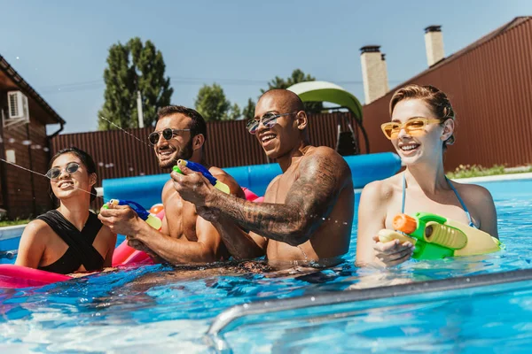Amigos multiétnicos felizes brincando com armas de água na piscina — Fotografia de Stock