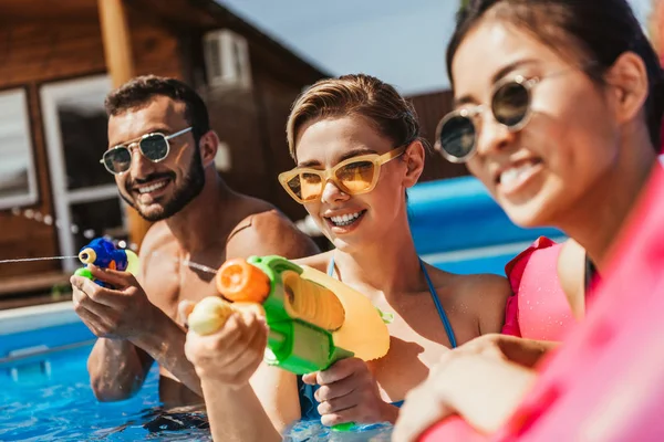 Pessoas multiétnicas em óculos de sol brincando com armas de água na piscina — Fotografia de Stock