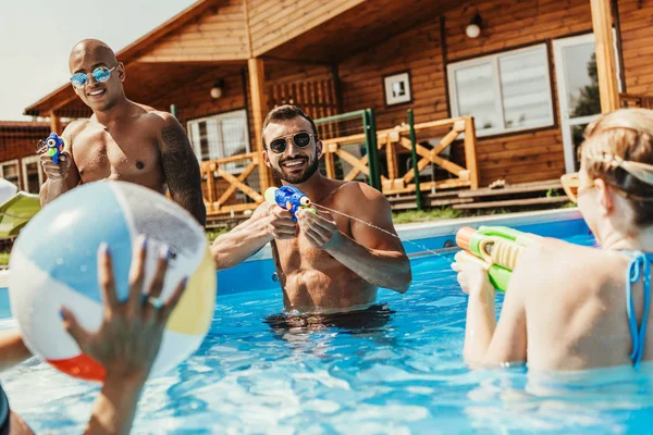 Amici multietnici che giocano con pallone da spiaggia e pistole ad acqua in piscina — Foto stock
