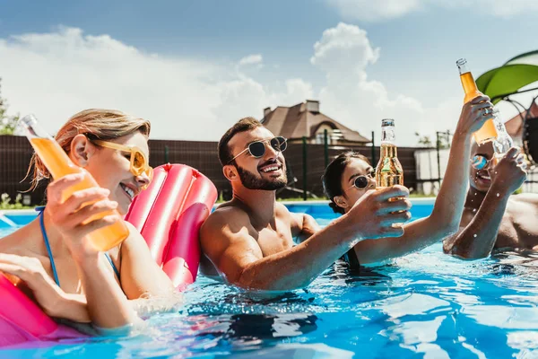 Multicultural friends with bottles of beer in swimming pool with inflatable mattress — Stock Photo