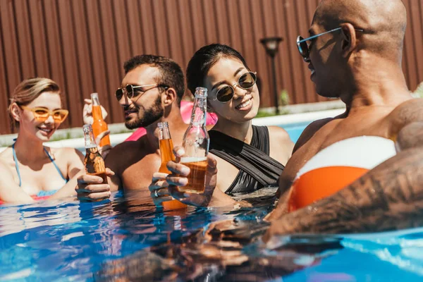 Amigos multiculturales con pelota de playa y botellas de cerveza en el agua en la piscina - foto de stock