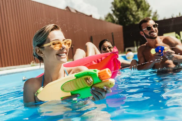 Amigos multiétnicos felices jugando con pistolas de agua en la piscina — Stock Photo