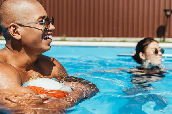 Afro-americano homem segurando praia bola na piscina com namorada atrás — Fotografia de Stock