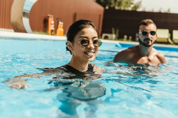 Attractive asian girl in swimming pool with boyfriend behind — Stock Photo