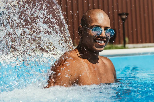 Усміхнений афроамериканський чоловік в сонцезахисних окулярах стоїть в басейні з бризками — стокове фото