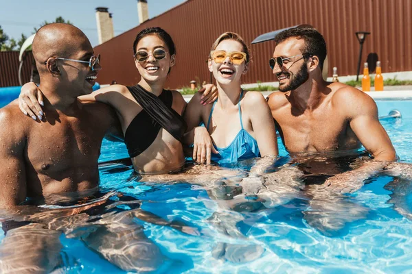 Rindo casais multiétnicos em óculos de sol passar tempo na piscina — Fotografia de Stock
