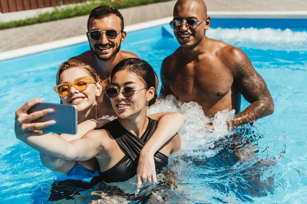 Multiethnic friends in sunglasses taking selfie on smartphone in swimming pool — Stock Photo