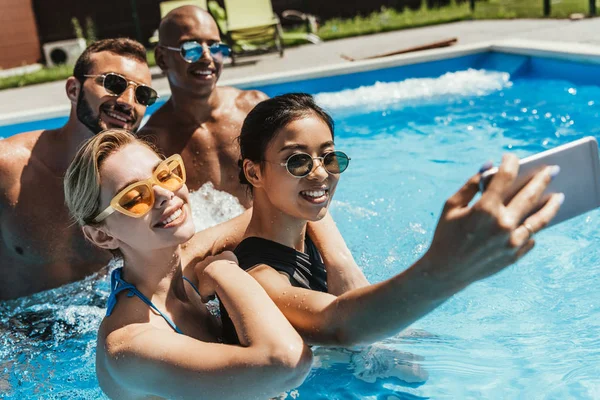 Parejas multiculturales en gafas de sol tomando selfie en smartphone en piscina — Stock Photo