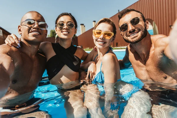 Amigos multiétnicos felices en trajes de baño y gafas de sol posando en la piscina - foto de stock