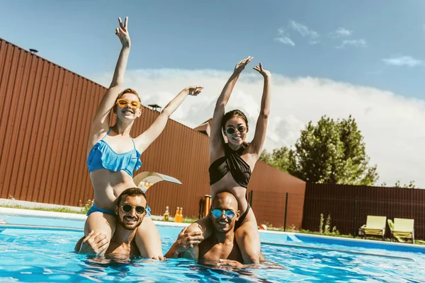 Happy beautiful multiethnic girls sitting on shoulders of boyfriends in swimming pool — Stock Photo