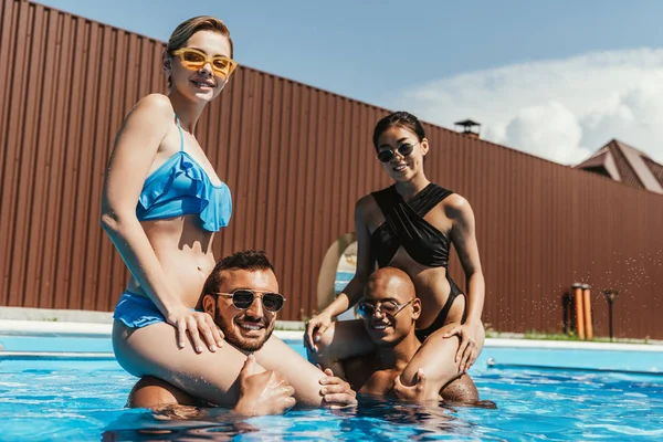 Multiethnic girls sitting on shoulders of boyfriends in swimming pool — Stock Photo