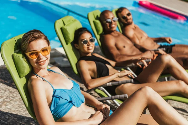 Smiling multicultural friends in swimsuits sunbathing on sunbeds at swimming pool — Stock Photo