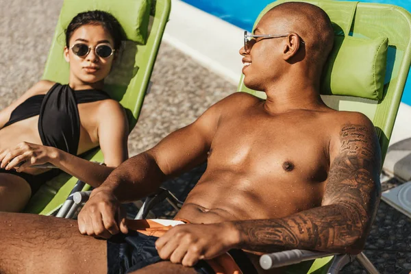 Casal interracial em óculos de sol relaxante em espreguiçadeiras à beira da piscina — Fotografia de Stock