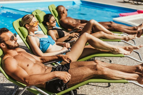 Amigos multiculturales en trajes de baño tomar el sol en tumbonas junto a la piscina, enfoque selectivo - foto de stock