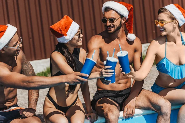 Багатоетнічні друзі в капелюхах Санти з напоями, що сидять біля басейну — стокове фото