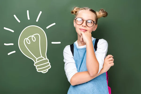 Pensive schoolchild in glasses looking up near blackboard with light bulb sign — Stock Photo