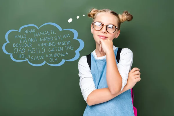 Pensive schoolchild in glasses looking up near blackboard with words on different languages in speech bubble — Stock Photo