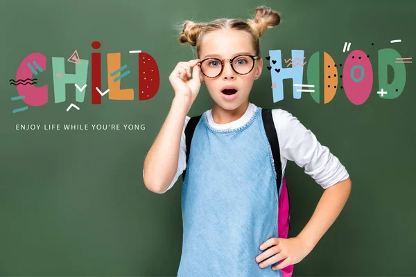 Shocked schoolchild touching glasses and looking at camera near blackboard, with 