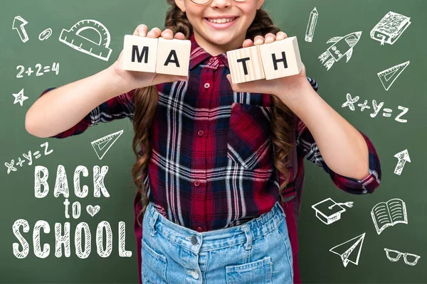 Imagem cortada de criança em idade escolar segurando cubos de madeira com palavra matemática perto de quadro negro com volta para a escola lettering — Fotografia de Stock