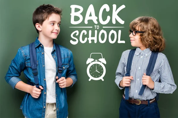 Schoolboys with backpacks looking at each other near blackboard, with alarm clock and 