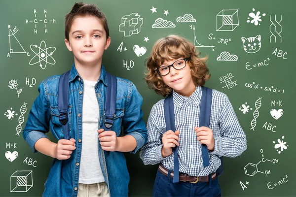 Schoolboys with backpacks looking at camera near blackboard with educational icons — Stock Photo