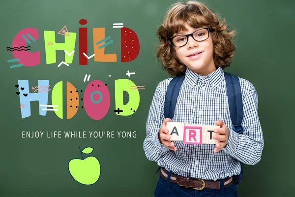 Schoolboy holding wooden cubes with word art near blackboard, with 