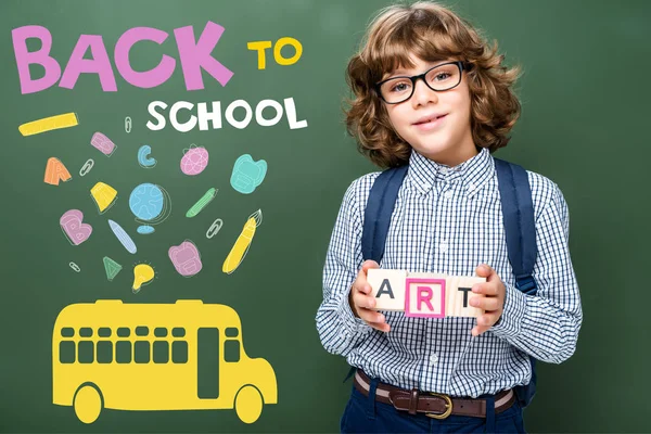 Schoolboy holding wooden cubes with word art near blackboard, with icons, bus and 