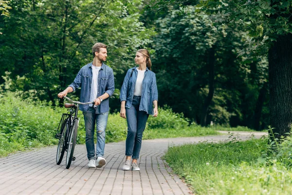 Beautiful young couple walking by park with vintage bicycle and looking at each other — Stock Photo