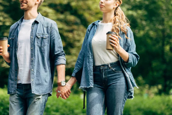 Plan recadré de jeune couple avec des tasses en papier de café marchant par parc ensemble — Photo de stock