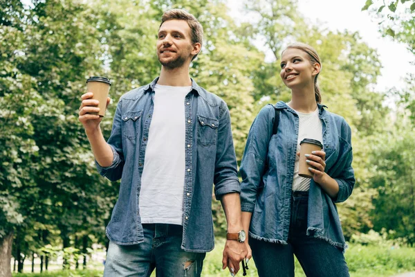 Blick von unten auf ein glückliches junges Paar mit Pappbechern Kaffee, das gemeinsam im Park spaziert — Stockfoto