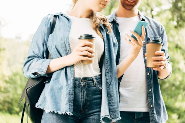 Recortado disparo de pareja joven con vasos de papel usando teléfono inteligente juntos en el parque - foto de stock