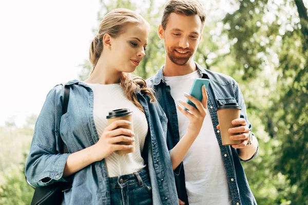 Hermosa pareja joven con vasos de papel utilizando el teléfono inteligente juntos en el parque - foto de stock