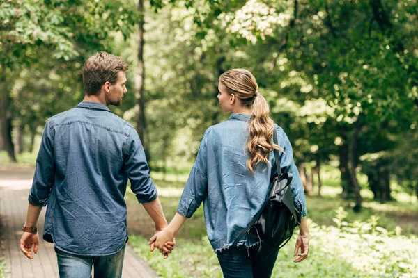 Vista trasera de hermosa pareja joven en camisas de mezclilla caminando por el parque - foto de stock