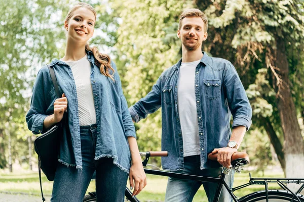 Heureux jeune couple avec vélo vintage debout au parc et regardant la caméra — Photo de stock
