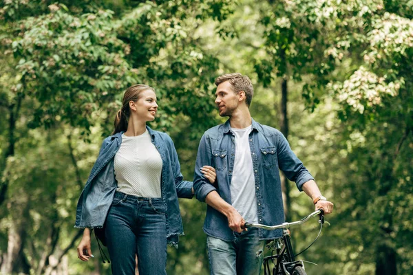 Heureux jeune couple en jeans chemises avec vélo marche par parc — Photo de stock