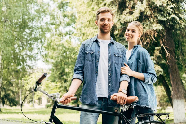 Belo jovem casal com bicicleta vintage de pé no parque e olhando para a câmera — Fotografia de Stock