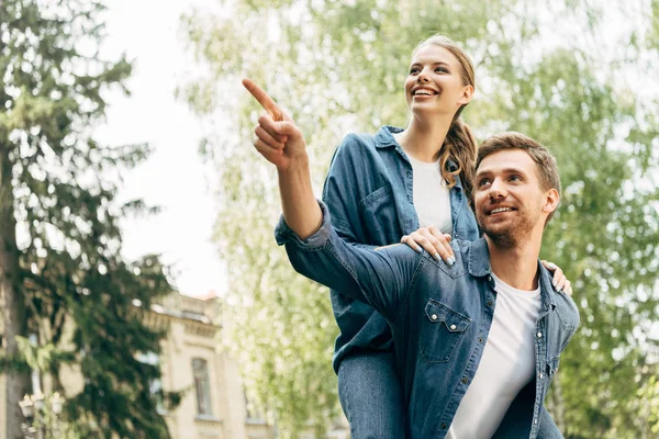 Hermosa joven mujer piggybacking en su novio mientras él señalando en algún lugar en el parque - foto de stock