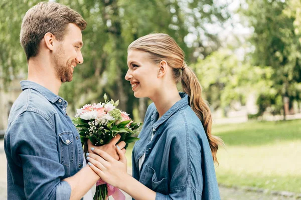 Felice giovane coppia che tiene mazzo di fiori al parco e si guarda a vicenda — Foto stock